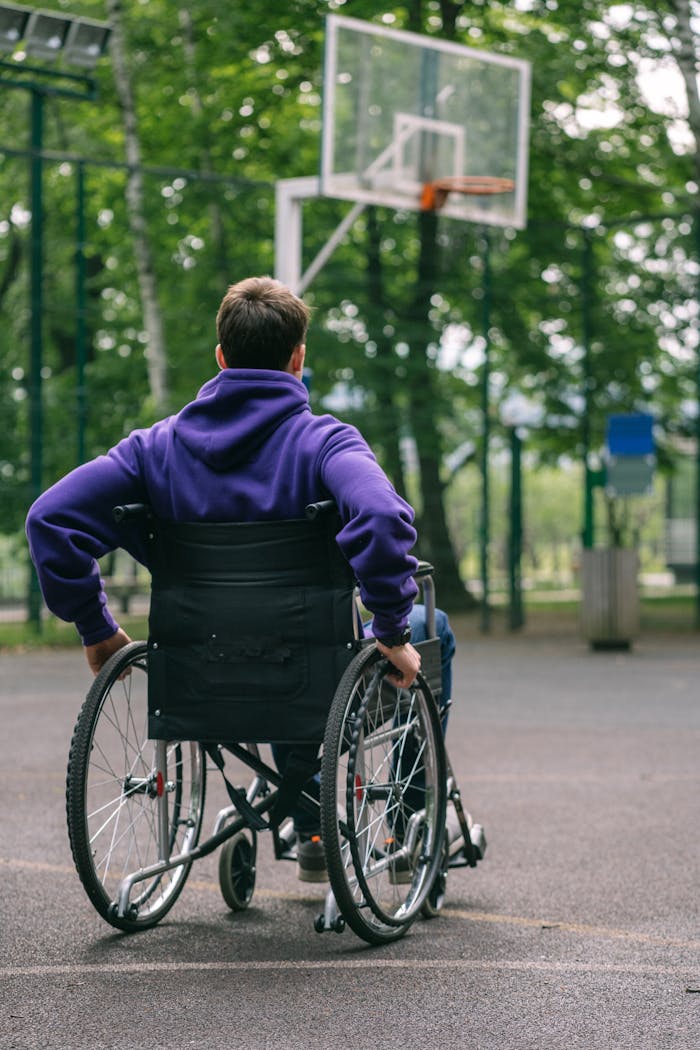 services-04 A man in a hoodie plays basketball on an outdoor court from a wheelchair, promoting inclusivity.